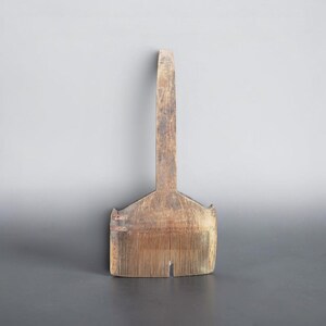 Puede incluir: Un peine de madera de color marrón claro desgastado con un mango vertical largo que se extiende desde una base rectangular ancha. La base contiene numerosos dientes finos y muy juntos, mostrando signos visibles de antigüedad y desgaste.
