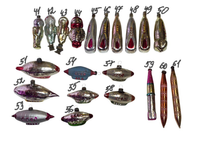 May include: A collection of vintage glass Christmas ornaments shaped like cosmonauts, rockets, and airships, numbered 41 through 61. The ornaments feature metallic finishes in silver, red, and blue, with some displaying the Cyrillic text CCCP.