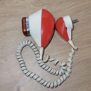 Peut inclure: Sèche-cheveux vintage avec un design rouge et blanc. Il est équipé d'un cordon blanc enroulé et d'une prise. L'ensemble est posé sur une surface en bois.