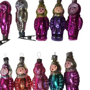 May include: A collection of ten vintage glass ornaments shaped like cosmonauts or figures in space suits. The ornaments feature metallic finishes in shades of pink, purple, silver, gold, and teal, each with a small metal loop for hanging.