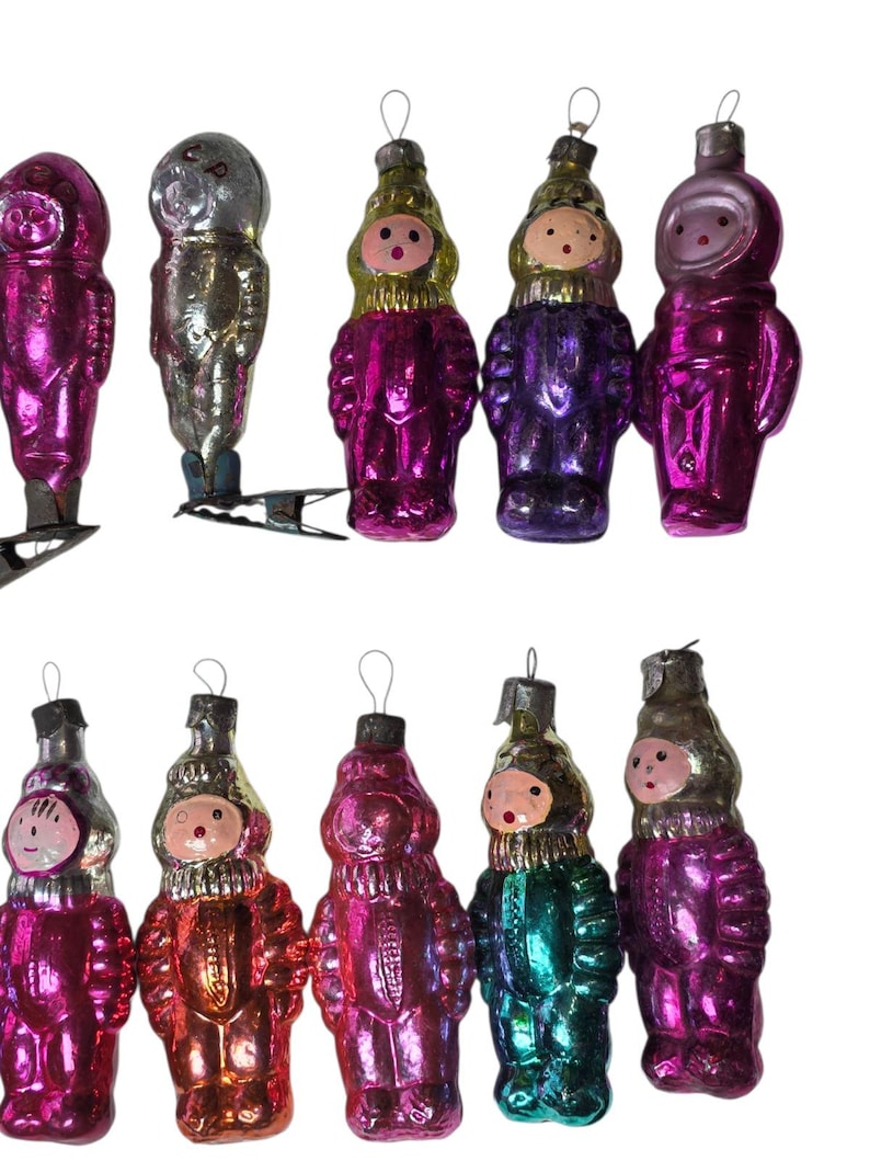 May include: A collection of ten vintage glass ornaments shaped like cosmonauts or figures in space suits. The ornaments feature metallic finishes in shades of pink, purple, silver, gold, and teal, each with a small metal loop for hanging.