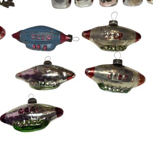 May include: A collection of vintage Christmas ornaments. The ornaments include a blue spaceship with the text "CCCP", and several silver and red UFO-shaped ornaments. The ornaments have a metallic finish and are designed for hanging on a Christmas tree.