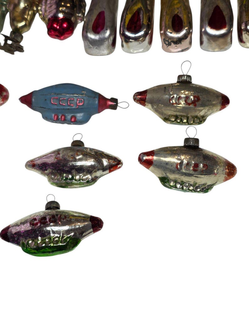 May include: A collection of vintage Christmas ornaments. The ornaments include a blue spaceship with the text "CCCP", and several silver and red UFO-shaped ornaments. The ornaments have a metallic finish and are designed for hanging on a Christmas tree.