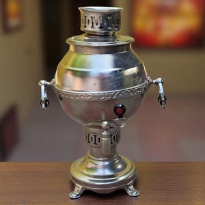 Può includere: Un samovar color argento con corpo arrotondato, rubinetto e maniglie decorative. Il samovar ha una parte superiore cilindrica e poggia su una base decorativa. Un pomello rosso è visibile sul lato. Il samovar è esposto su una superficie di legno.