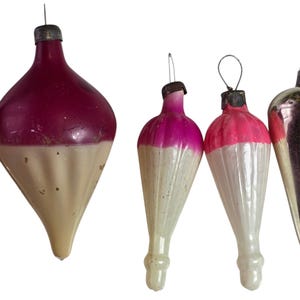 May include: A collection of four vintage glass ornaments featuring a two-tone design with a vibrant pink or magenta upper section and a cream-colored, ribbed lower section. Each ornament is topped with a metallic cap and a wire loop.