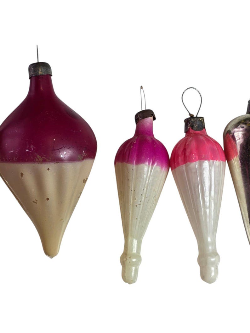 May include: A collection of four vintage glass ornaments featuring a two-tone design with a vibrant pink or magenta upper section and a cream-colored, ribbed lower section. Each ornament is topped with a metallic cap and a wire loop.