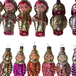 May include: A collection of vintage Christmas ornaments in the shape of figures. The ornaments have a metallic finish in gold, silver, pink, and red. Each ornament features a face and is wearing a hooded outfit.