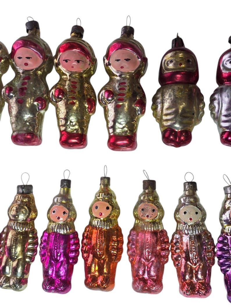 May include: A collection of vintage Christmas ornaments in the shape of figures. The ornaments have a metallic finish in gold, silver, pink, and red. Each ornament features a face and is wearing a hooded outfit.