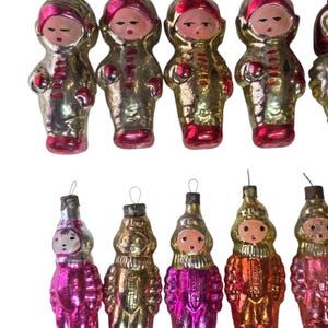 May include: A collection of vintage Christmas ornaments shaped like figures in space suits. The ornaments are primarily gold with red and pink accents, featuring painted faces and silver hanging loops.