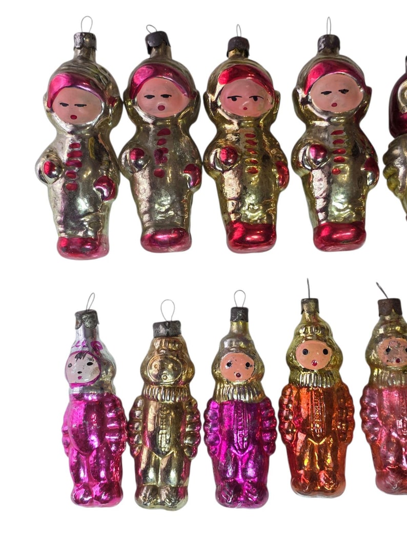 May include: A collection of vintage Christmas ornaments shaped like figures in space suits. The ornaments are primarily gold with red and pink accents, featuring painted faces and silver hanging loops.