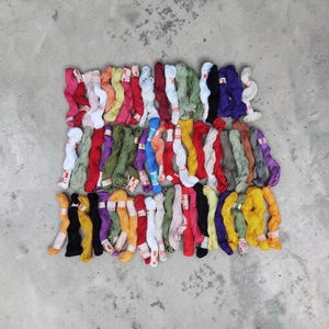 May include: A collection of embroidery floss skeins arranged in three horizontal rows. The threads feature a wide variety of solid colors including red, blue, green, yellow, pink, purple, orange, black, white, and brown, each with a paper label.