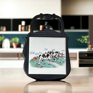 Może przedstawiać: Czarny żeliwny podkładka w kształcie czajnika z wkładką z białej ceramiki przedstawiającą kreskówkową ilustrację dwóch czarno-białych krów na łące z czerwonym stodołą i niebieskim niebem.