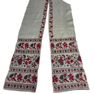 May include: A long, off-white scarf with intricate floral embroidery in red, black, and brown. The embroidery features a repeating pattern of flowers and leaves along the edges and ends of the scarf. The scarf is made of a woven fabric.