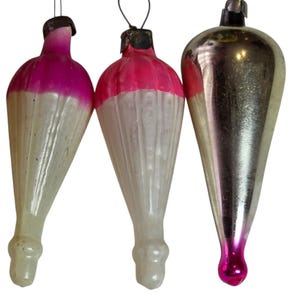 May include: Three vintage Christmas ornaments. Two are white with pink tops, and the third is silver with a pink base. Each ornament has a metal loop for hanging. The ornaments are teardrop-shaped.