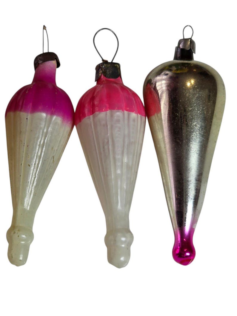 May include: Three vintage Christmas ornaments. Two are white with pink tops, and the third is silver with a pink base. Each ornament has a metal loop for hanging. The ornaments are teardrop-shaped.