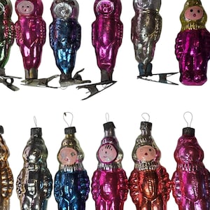May include: A collection of vintage Christmas ornaments in various colors, including pink, blue, and silver. The ornaments are shaped like figures with faces and are attached to metal clips for hanging on a tree.