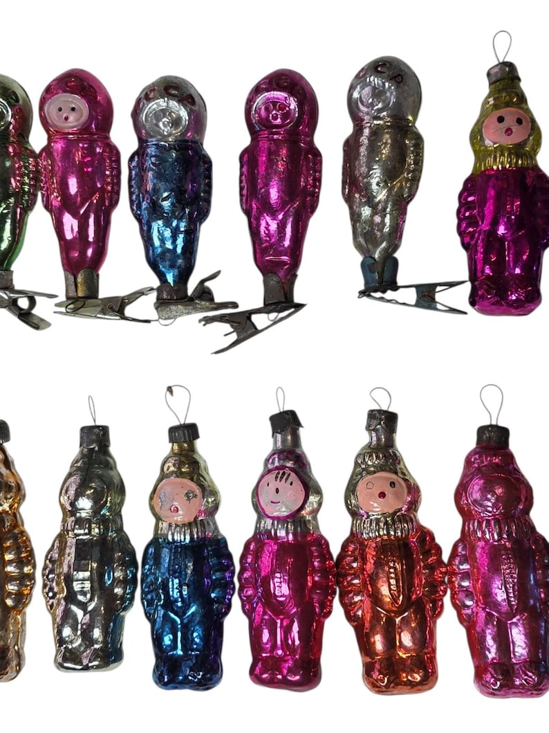 May include: A collection of vintage Christmas ornaments in various colors, including pink, blue, and silver. The ornaments are shaped like figures with faces and are attached to metal clips for hanging on a tree.