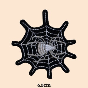 Op de afbeelding: Zwart-wit geborduurde strijkpatch van een spin in een web. De spin is wit en het web is zwart. De patch heeft een diameter van 6,5 cm.