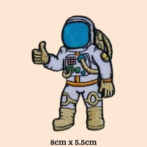 Puede incluir: Un parche bordado para planchar de un astronauta dando un pulgar hacia arriba. El astronauta lleva un traje espacial blanco con una visera azul y detalles naranjas. El parche mide 8 cm por 5,5 cm.