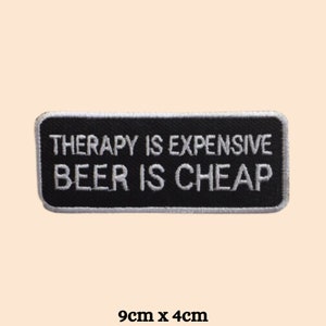 Op de afbeelding: Zwart-witte geborduurde patch met de tekst "Therapy is expensive, Beer is cheap" op een witte achtergrond. De patch is 9 cm x 4 cm.