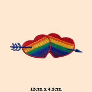 Könnte beinhalten: Zwei regenbogenfarbene Herzen mit einem Pfeil, der sie durchstößt, symbolisieren Liebe und Stolz. Die Herzen sind rot mit Regenbogenstreifen und der Pfeil ist blau. Der Patch misst 12 cm x 4,2 cm.