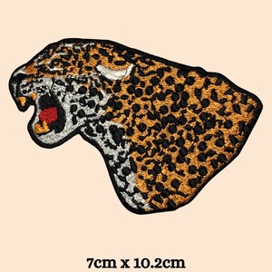 Może przedstawiać: Naszywka z haftem przedstawiająca ryczącą głowę jaguara w kolorze czarnym i pomarańczowym. Naszywka ma wymiary 7 cm x 10,2 cm.