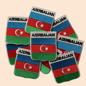 May include: A stack of embroidered patches featuring the flag of Azerbaijan. Each patch is rectangular with a white border and the words "AZERBAIJAN" printed in black. The flag is blue, red, and green with a white crescent moon and star in the center.