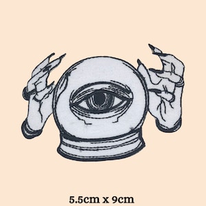 Op de afbeelding: Geborduurde patch met een kristallen bol met een oog en handen. Het ontwerp is in zwart en wit, met de kristallen bol van ongeveer 5,5 cm bij 9 cm. De patch heeft een mystiek of occult thema.