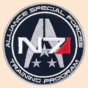 May include: A circular patch with a dark blue background and white text. The text reads "Alliance Special Forces Training Program". In the center of the patch is a white "N7" logo with a red triangle. The logo is surrounded by three white stars.