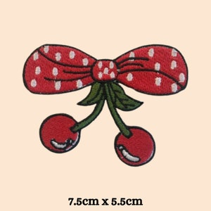 Pode incluir: Laço vermelho com bolinhas brancas e duas cerejas vermelhas presas ao laço. As cerejas têm hastes e folhas verdes. O laço mede 7,5 cm por 5,5 cm.