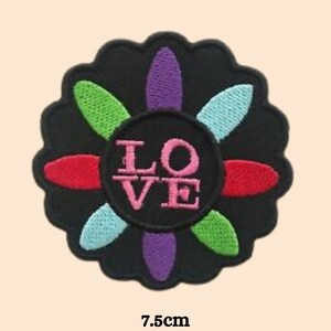 Puede incluir: Un parche bordado negro en forma de flor con seis pétalos. Los pétalos son de diferentes colores: rojo, verde, azul, morado. En el centro de la flor hay un círculo con la palabra "LOVE" en letras rosas.