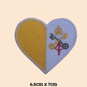 May include: An embroidered patch in the shape of a heart, depicting the Vatican City flag. The flag is yellow and white with the papal coat of arms on the white side. The patch measures 6.5 cm by 7 cm.