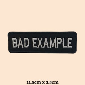 May include: Black embroidered patch with white text that reads "BAD EXAMPLE". The patch is 11.5 cm by 3.5 cm.