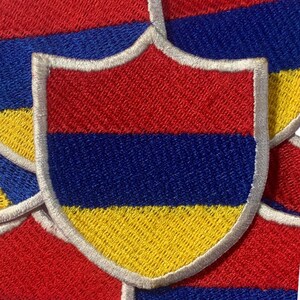 May include: A red, blue, and yellow embroidered patch in the shape of a shield. The patch has a white border.