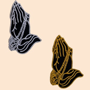 Peut inclure: Deux patchs brodés à repasser représentant des mains en prière avec une chaîne autour des poignets. Un patch est noir avec des coutures blanches et l'autre est noir avec des coutures dorées.