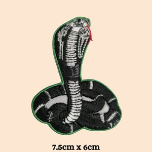 May include: Green and black embroidered iron-on patch of a cobra with its hood raised. The snake is white with black markings. The patch measures 7.5 inches by 6 inches.