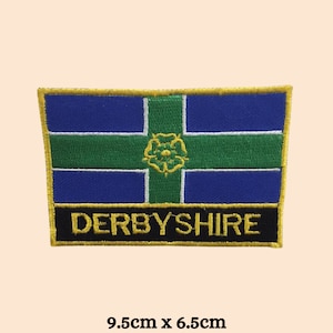 May include: A blue, green and white embroidered patch with a gold border and the text "DERBYSHIRE" in gold lettering. The patch is 9.5cm by 6.5cm.