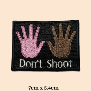 May include: Black and pink embroidered patch with two hands raised in the air and the text "Don't Shoot" in white. The patch is 7 cm by 5.4 cm.