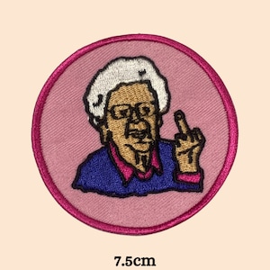 Op de afbeelding: Een geborduurde strijk-patch met een cartoon van een persoon met wit haar, die een bril en een blauwe trui draagt en de middelvinger opsteekt. De patch heeft een diameter van 7,5 cm.