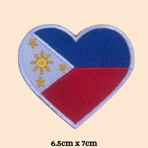 May include: A heart-shaped embroidered patch featuring the flag of the Philippines. The patch is white with a blue triangle at the hoist and a red triangle at the fly. A yellow sun with eight rays and three five-pointed stars are in the white triangle. The patch measures 6.5 inches by 7 inches.