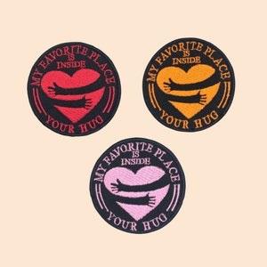 May include: Three embroidered patches with the text "MY FAVORITE PLACE IS INSIDE YOUR HUG". Each patch features a heart with hugging arms. The patches are in red, orange, and pink, with black borders.