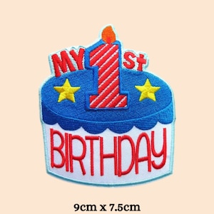 May include: A blue and white embroidered patch with a red and white striped candle on a cake. The cake has two yellow stars and the text "My 1st Birthday" in red. The patch is 9 cm by 7.5 cm.