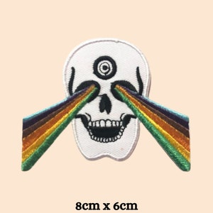 Könnte beinhalten: Weißer gestickter Patch mit einem Totenkopf, der ein drittes Auge hat und dessen Augenhöhlen Regenbogenstrahlen aussenden. Der Patch ist 8 cm x 6 cm groß.