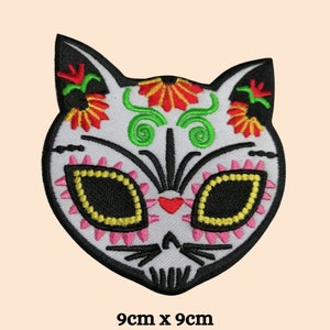May include: White embroidered iron-on patch of a cat with a Day of the Dead sugar skull design. The cat has black eyes with yellow accents, a pink nose, and green and red floral details. The patch measures 9 inches by 9 inches.