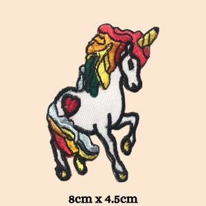 Pode incluir: Um unicórnio branco com uma crina e cauda arco-íris. O unicórnio tem um coração vermelho no lado e um chifre dourado. O remendo mede 8 cm por 4,5 cm.