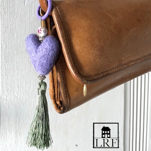 May include: A purple felt heart charm attached to a lavender plastic ring, featuring a white floral bead, silver-toned spacers, and a sage green thread tassel. The charm hangs from the edge of a brown leather bag with a gold-toned metal pull.