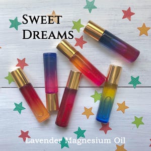 May include: Five cylindrical glass roller bottles with gold caps, featuring gradient colours of blue, pink, orange, and yellow. The text SWEET DREAMS and Lavender Magnesium Oil is printed on the white surface decorated with colourful paper stars.