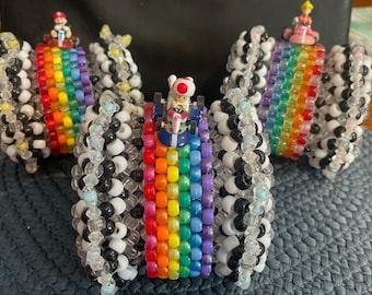 May include: Three colourful beaded bracelets with rainbow patterns and small toy figures on top. The bracelets are made with black, white, and rainbow coloured beads.
