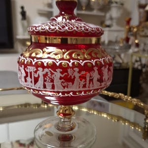 Peut inclure: Un sucrier en verre rouge rubis avec couvercle assorti, orné de figures blanches de style camée représentant des personnes en tenue classique, de volutes dorées peintes et d'une base en verre transparent avec un liseré doré sur le bol.