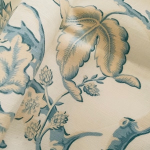 May include: Close-up of a fabric with a floral pattern. The design features large leaves and floral elements in shades of blue and beige against a cream background. The fabric is folded, showing the texture and depth of the material.